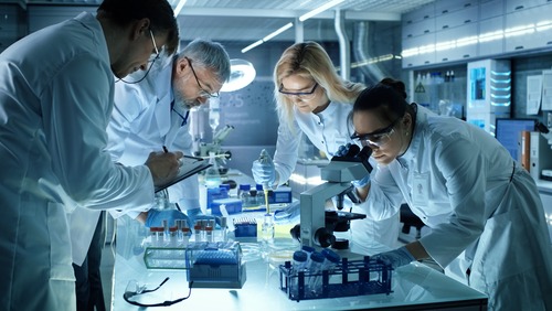 Four people in protective gear work hunched together over a table in a lab setting
