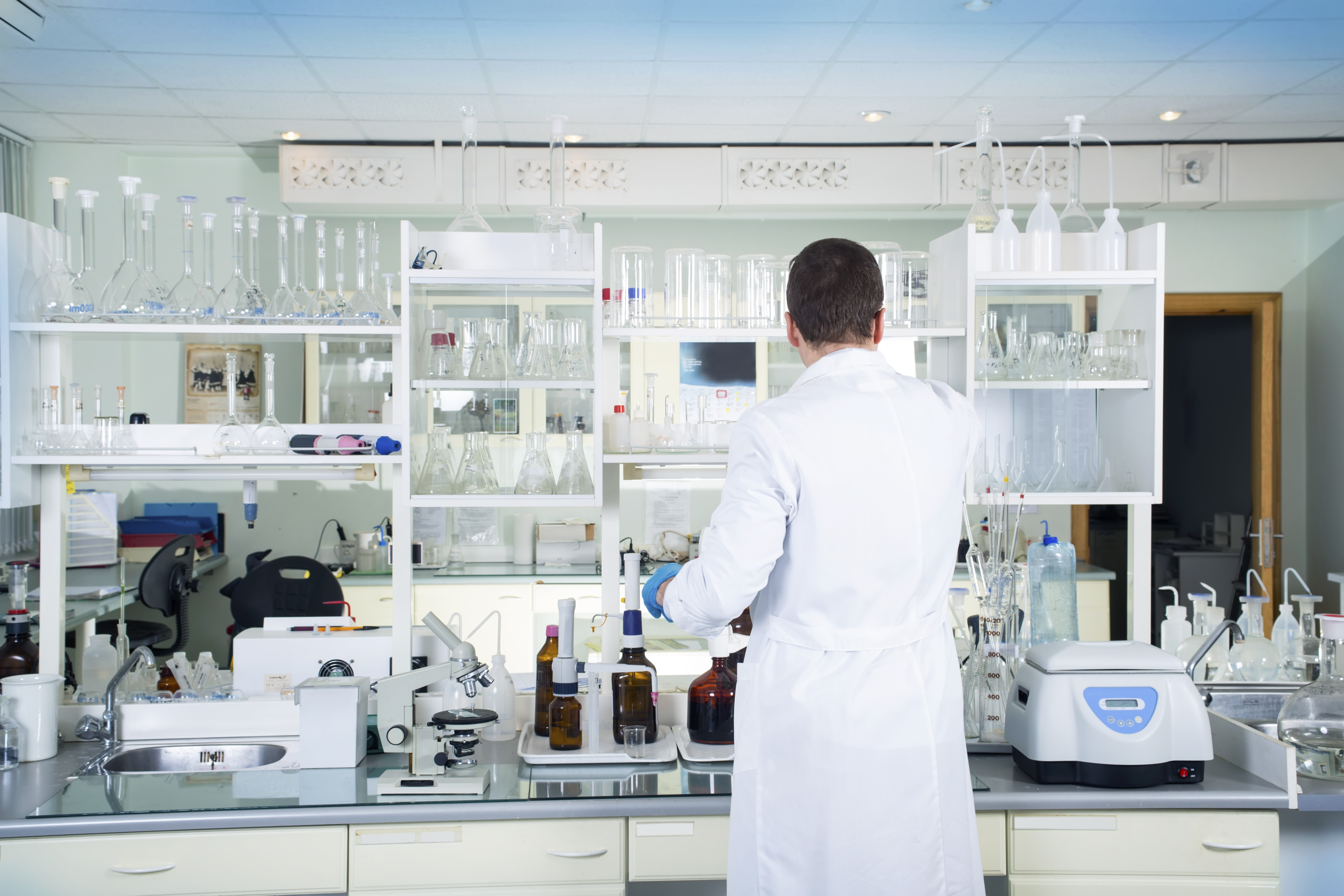 A person with dark hair in a white labcoat faces away from the camera in a laboratory setting